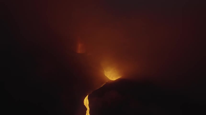 Orange glowing river of lava flows down a hillside from the mouth of the volcano at night