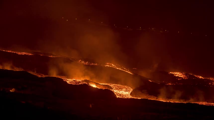 Time lapse of a smoke rising and a glowing orange river of lava flowing down a hillside at night
