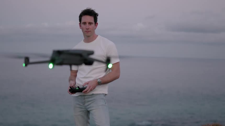 A man standing on a cliff above the sea pilots a drone making it hover in front of him