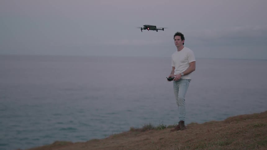 A man standing near the sea is ascending a drone using a controller