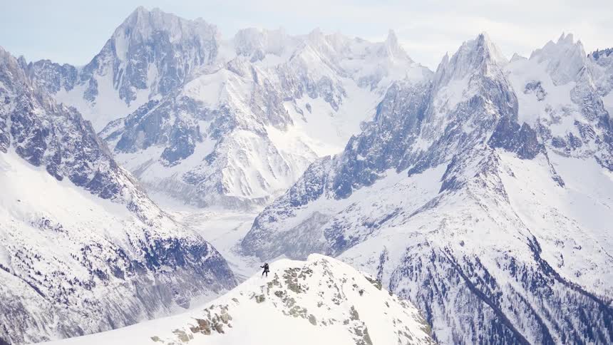 Skier stands at the top of a snow covered peak surrounded by jagged mountain peaks during the day