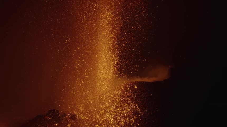 Orange lava sparks shoot up from the mouth of erupting volcano and land on a hillside at night