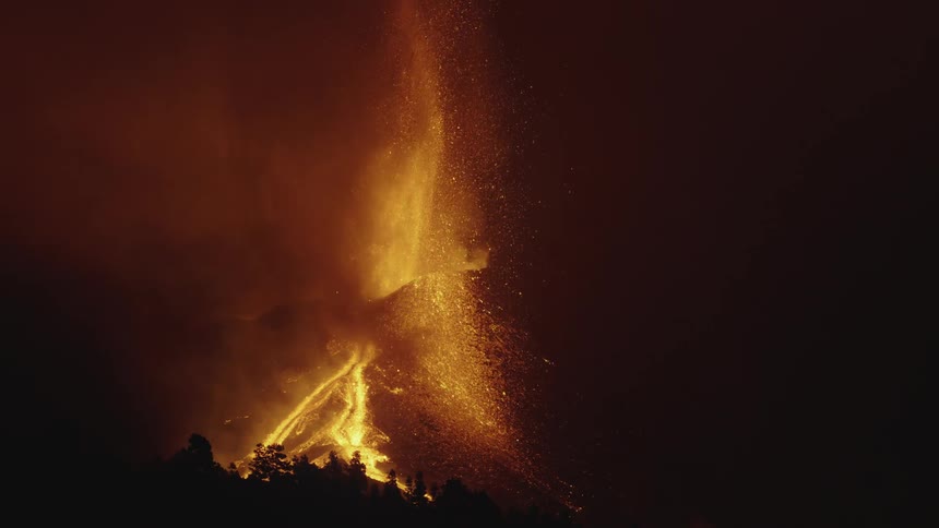 Volcano erupts with a fountain shooting up and a river of orange glowing lava flowing down the hillside at night in slow motion