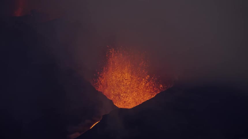Lava sprays into smoky air and flows down slope of an erupting volcano at night