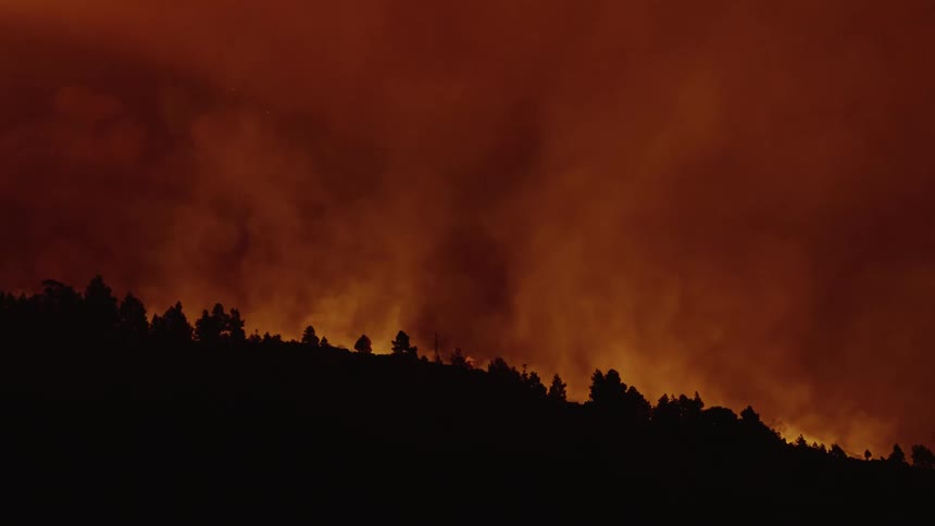 Smoke rises up and orange glow from behind a silhouetted tree line at night