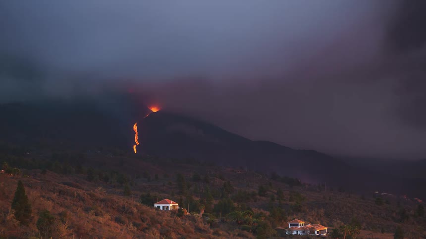 A volcano erupting on a mountain near some houses in a rural region