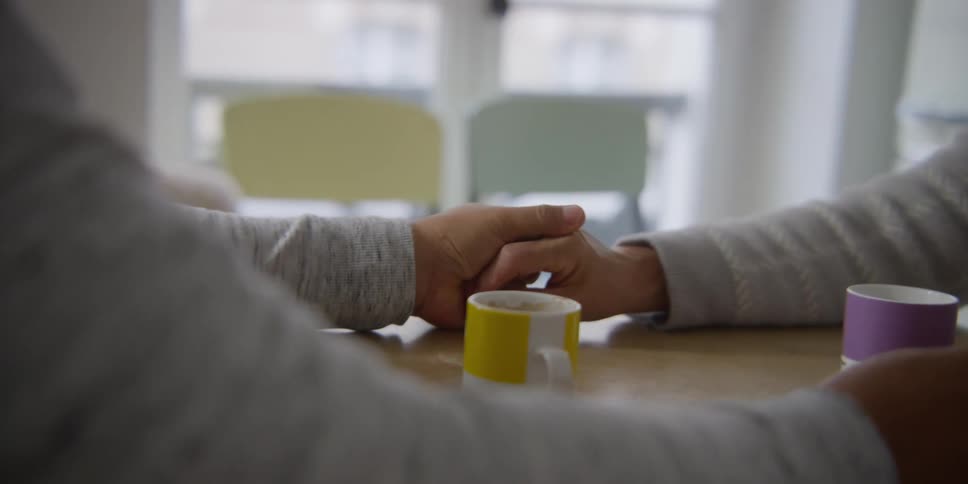 A couple holds each others hands over a table with two mugs in front of them