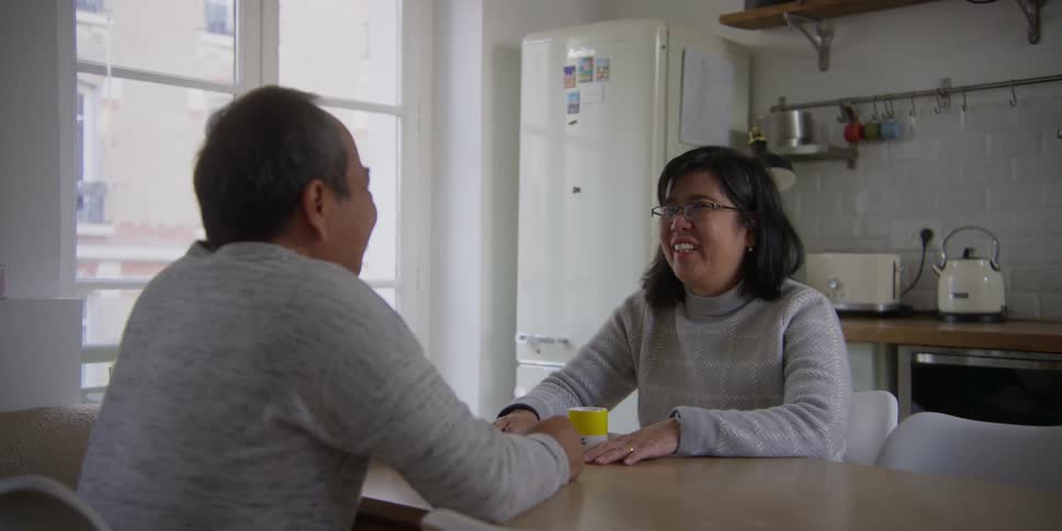 A couple sitting at a kitchen table smiling and laughing with two mugs in front of them