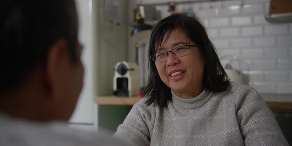 An asian woman wearing glasses is talking and smiling while talking to someone in front of her