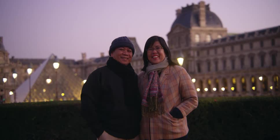 A couple is wearing warm clothes and posing for a photo and behind them is a large building