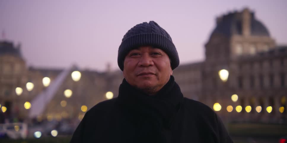 Older man in coat stands smiling as traffic passes between him and large building in the background