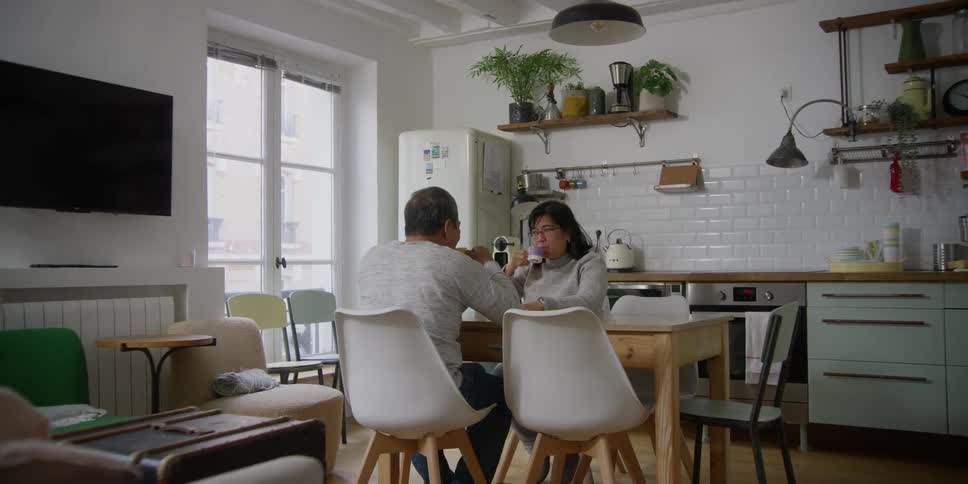 Older couple sits at table near kitchen drinking from mugs