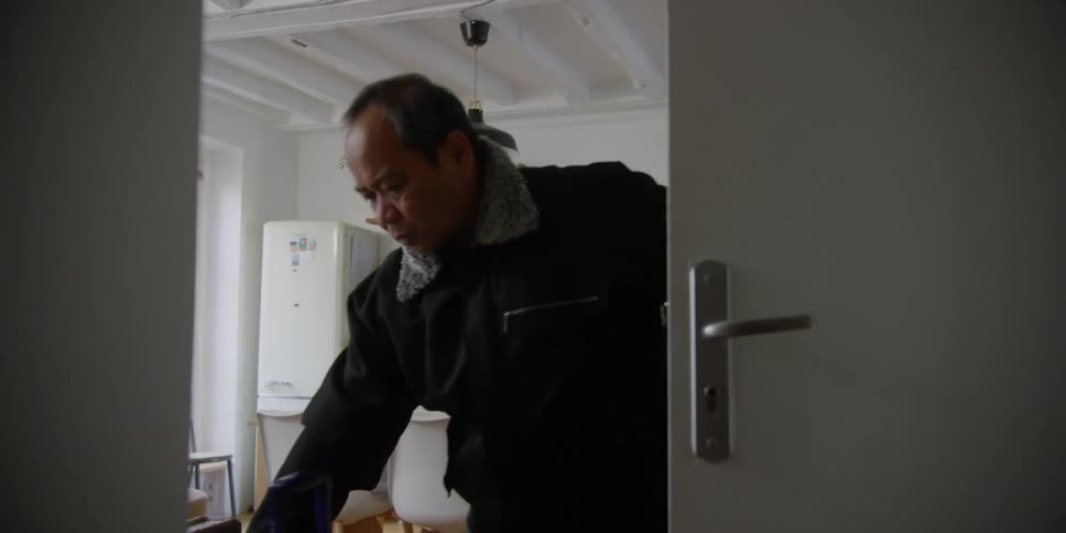 A man and a woman in warm clothes enter through a white door into a room that has a kitchen