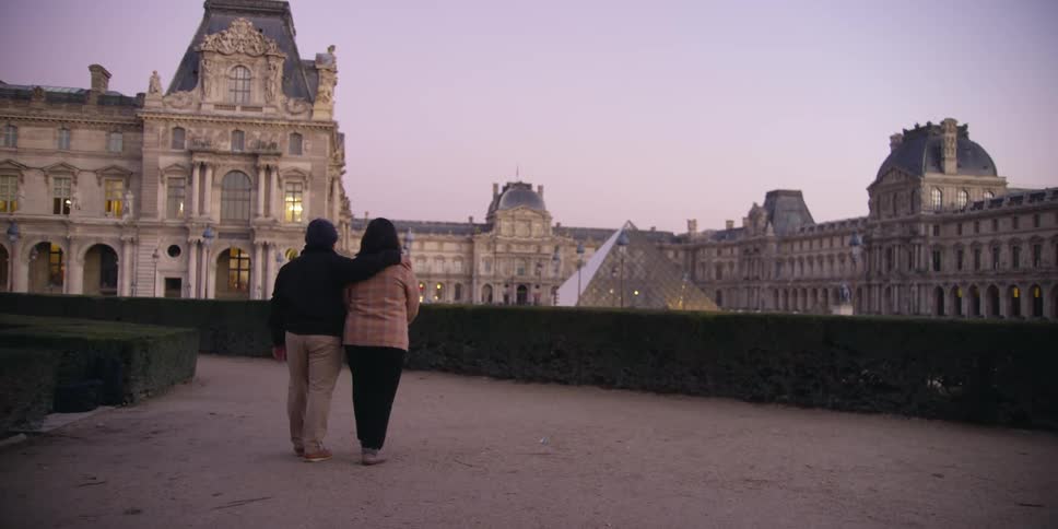 Couple out for a casual evening stroll on the rooftop of one of the buildings of The Louvre Museum in Paris, France as they view the glass pyramid and beautiful architecture around them..