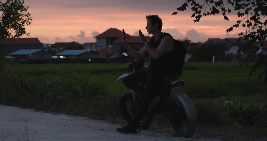 Man waves during video call while sitting on motorcycle at side of road with houses in the background