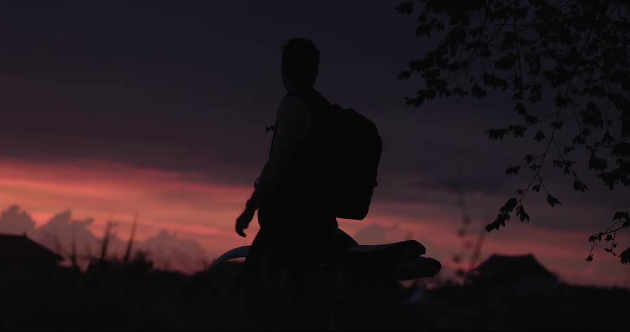 Man looking at his smartphone while sitting on motorcycle stands partially silhouetted against a red sky
