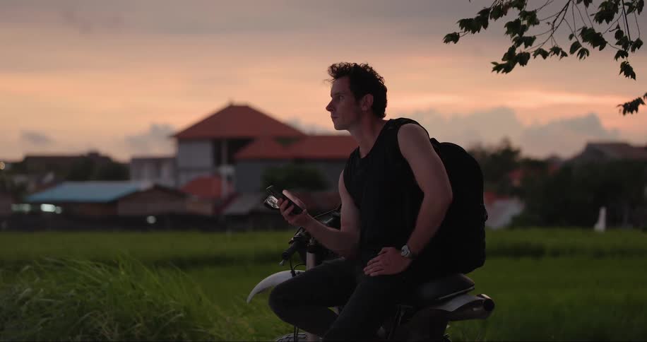 A man looking down on his mobile phone while seated on a motorcycle outdoor in a grass field at sunset
