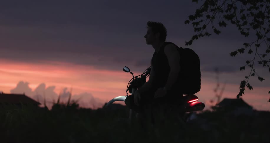 A man sits atop his idle dirt bike while looking at his phone at dusk