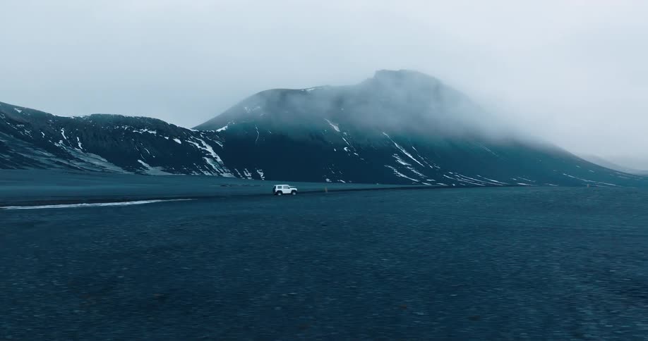 Vehicle drives through plain bordered by snow dusted mountains on a foggy day