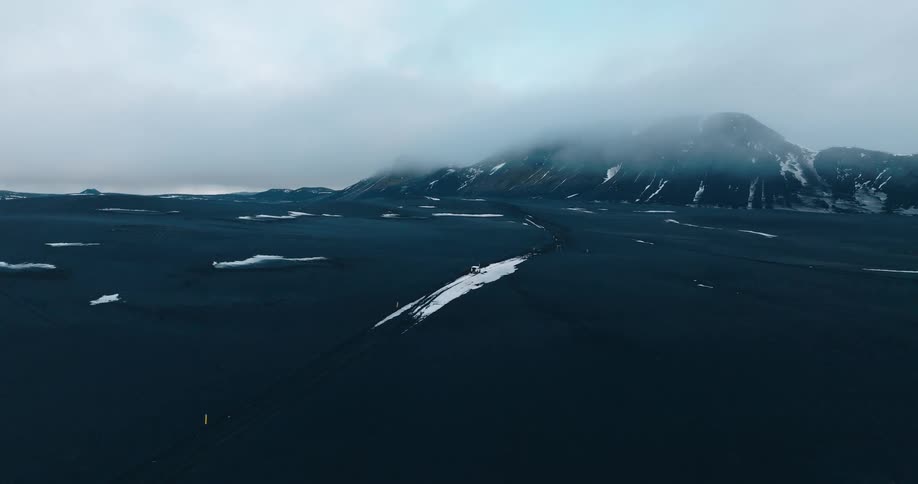 Aerial view of an off road vehicle on a desolate landscape with snow and mountains under a cloudy sky