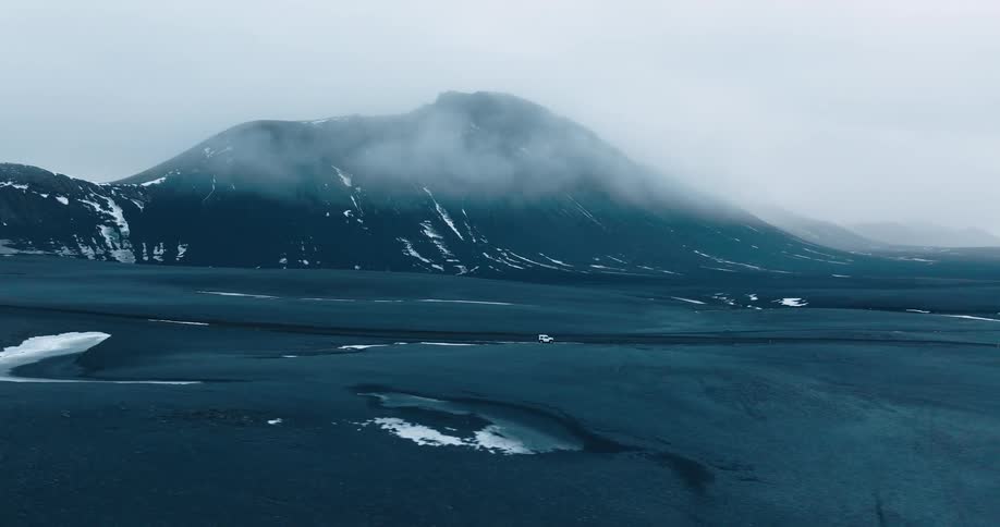 A white off road vehicle drives through a desolate landscape with snow and mountains under a cloudy sky
