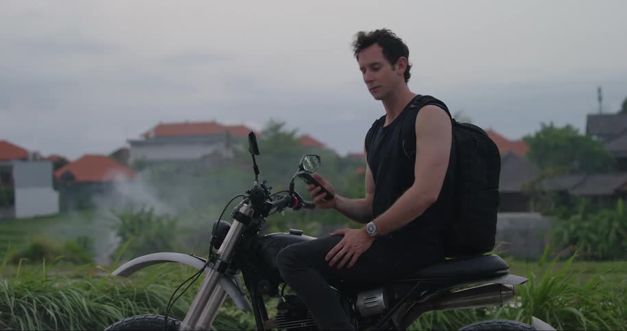 A white man on a motorcycle looks around as other people drive by him