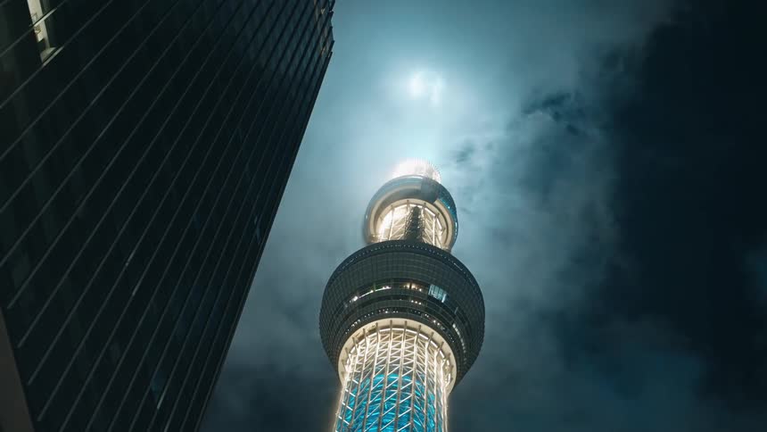 Brightly lit space needle illuminates clouds passing over nearby skyscraper at night