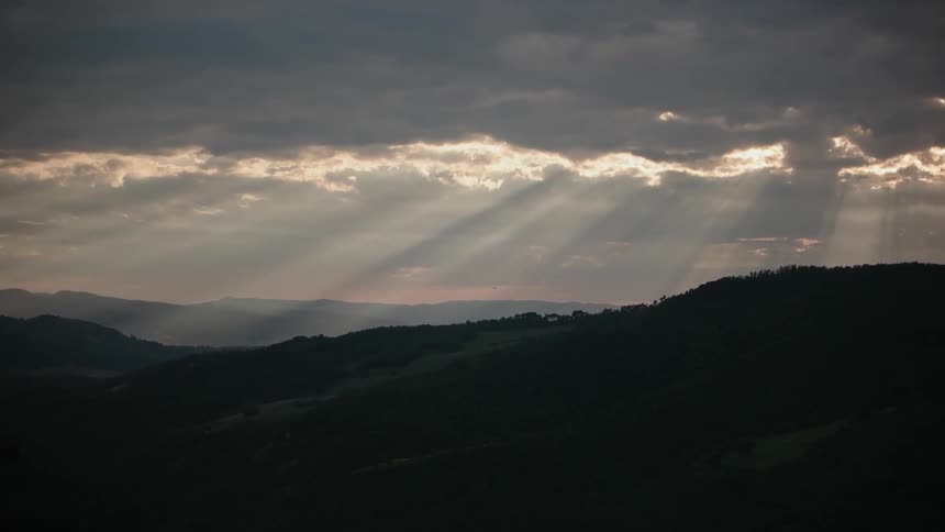 Rays of light shining through an opening in the clouds and shining down on a hilly area