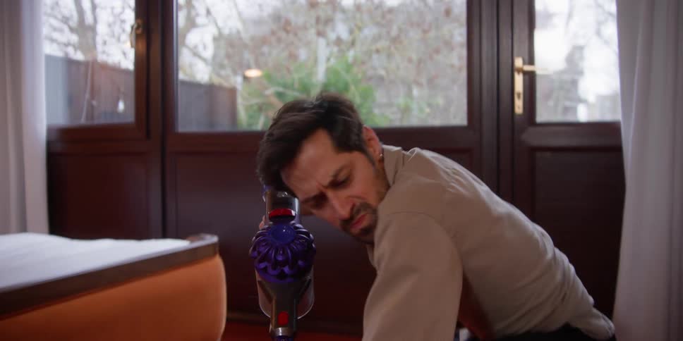 A man using a cordless vacuum cleaning in a bedroom playfully dances