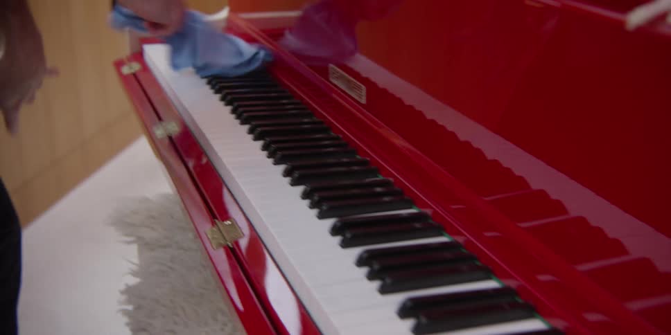 A man is cleaning a red piano using a blue cloth and then blows on the piano keys