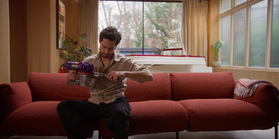 A man grabs a small sweeper and cleans the couch