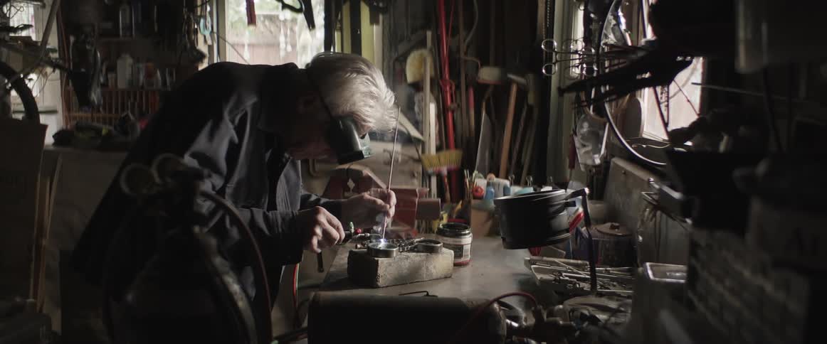 Older man solders item on workbench in garage