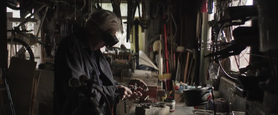 An old man dons goggles and lights a torch in a workshop
