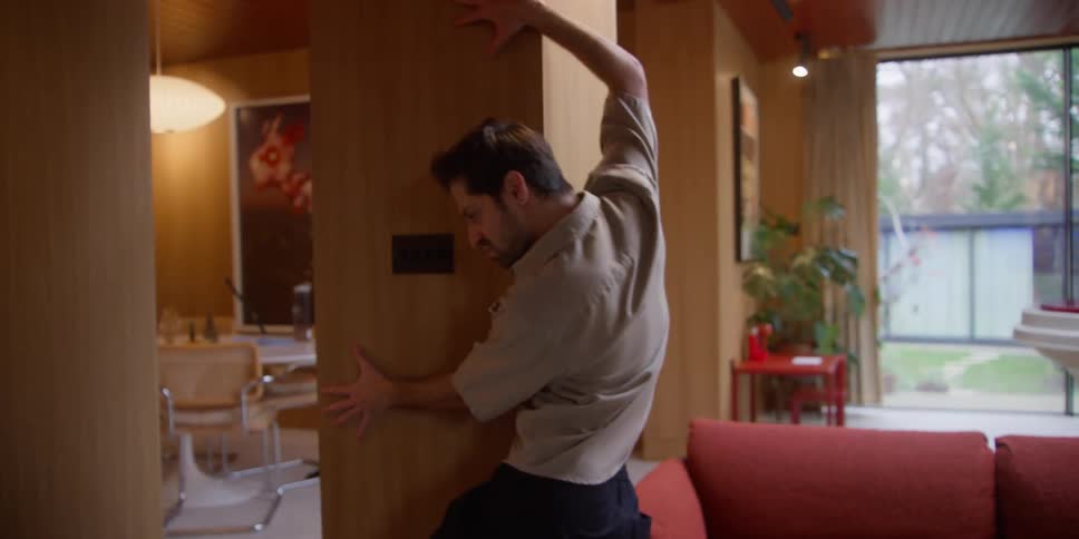 A man is doing an artistic performance inside a living room where he is holding onto the bookshelf and then goes to the sofa