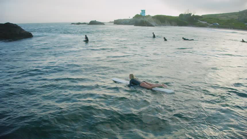 A region of the sea with several surfers and there is a woman on the board paddling and then sitting on the board