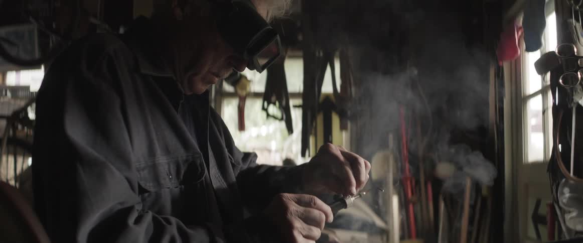 An old man is wearing glasses and using a blowtorch on something and smoke is coming out