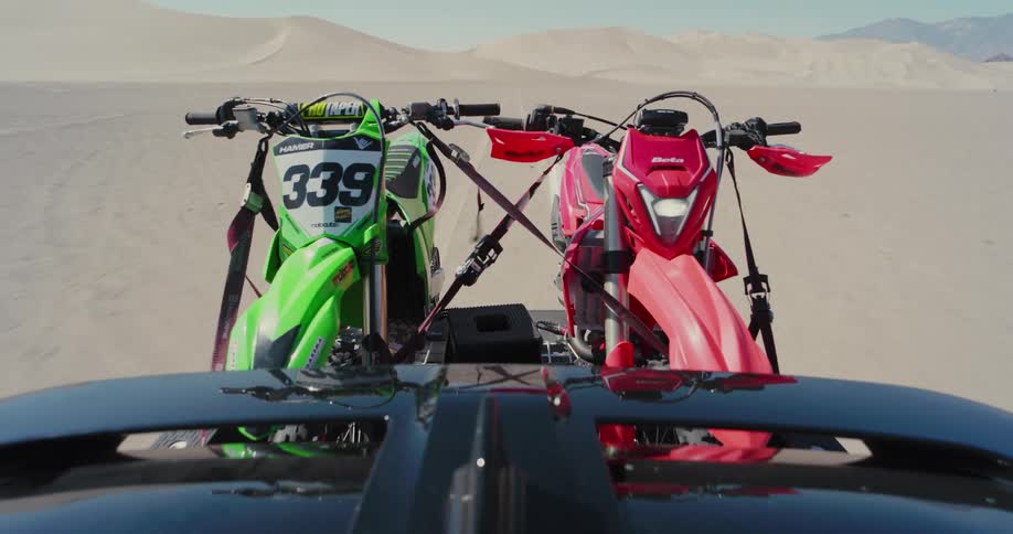 A green and a red motorcycle are tied next to each other and being pulled through a desert