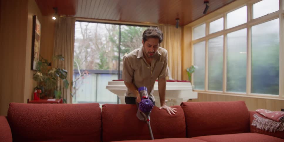 A man dances and rolls over a couch while cleaning it with a cordless vacuum