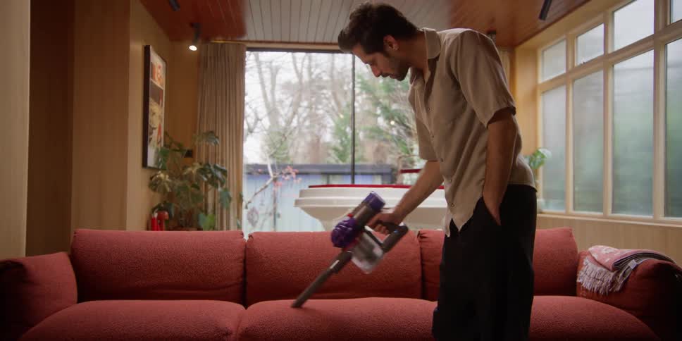 A man sitting on the sofa gets up and then starts vacuuming the sofa with a vacuum cleaner