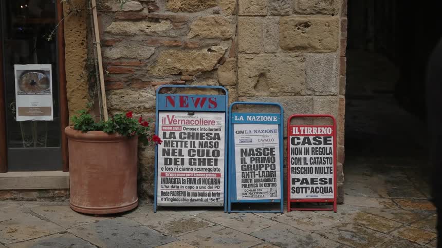 People walk in front of news signs in front of a stone building