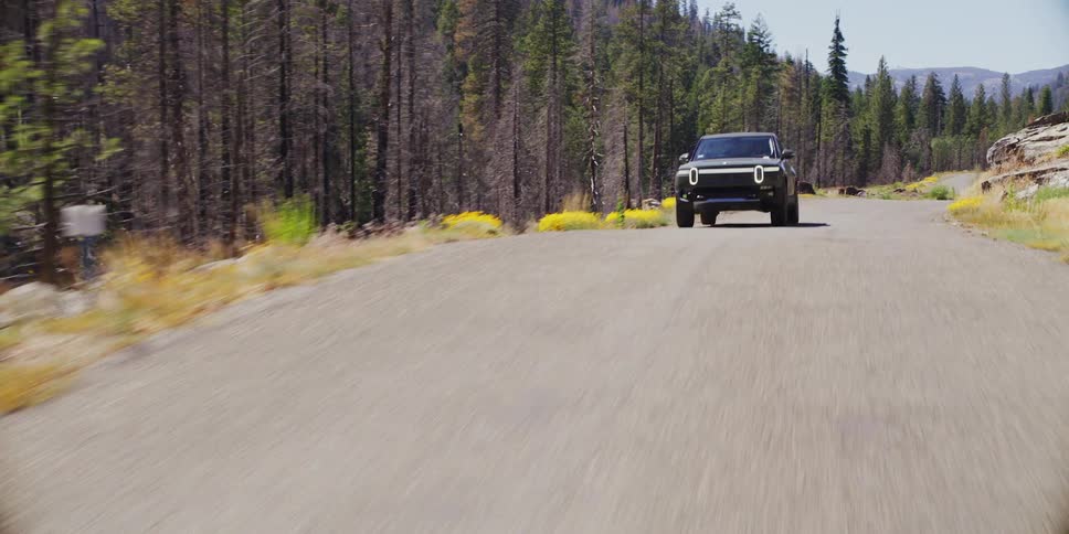 A car is driving on a rural road near a pine forest and several rocks on the side of the road