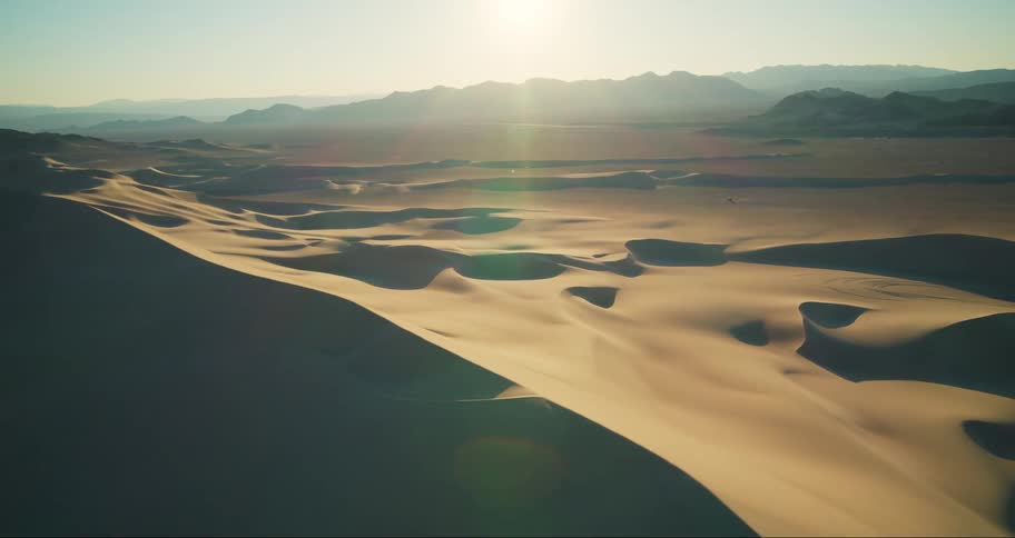 Bright sun shines down onto desert dunes from above nearby foggy mountains