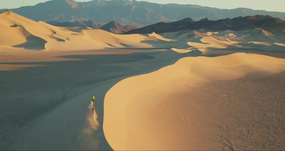 Two motorcyclists ride across a sand dune in a desert