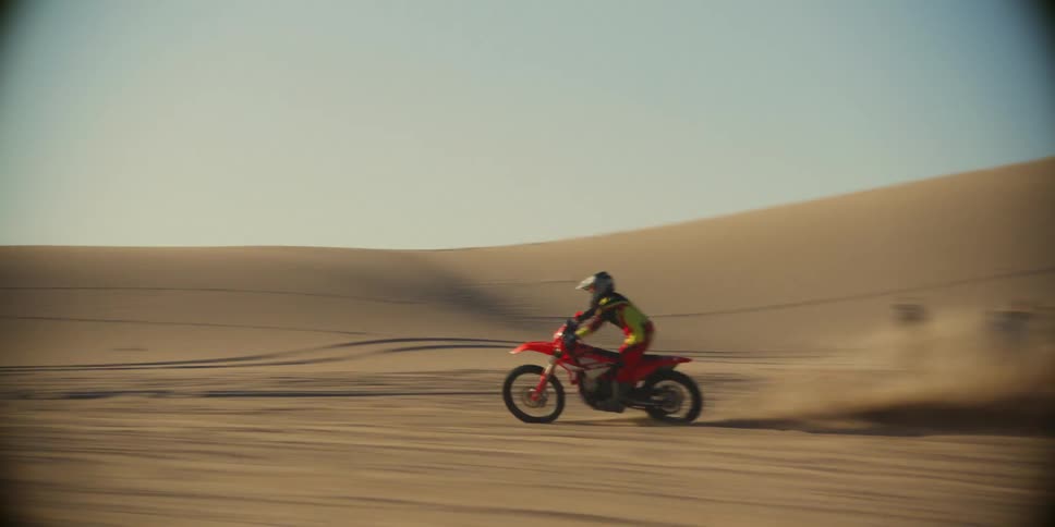 Two motorbikes and a car are driving in a desert and kicking up a lot of sand and one of the motorbikes rears up