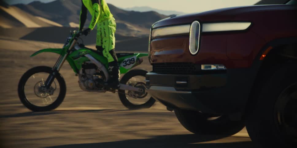 Man on a motorcycle next to a red pickup truck both traveling in the middle of the desert