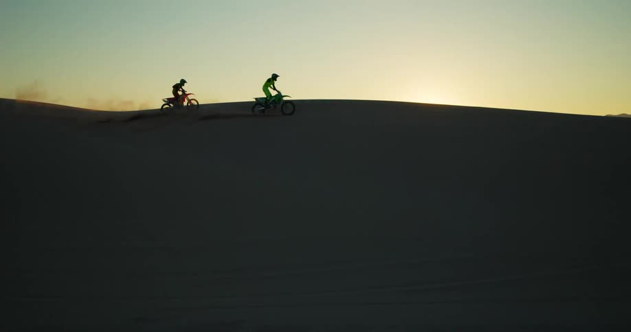 Two people riding motocross at high speed through the desert dunes and a late afternoon with a sunset on the horizon