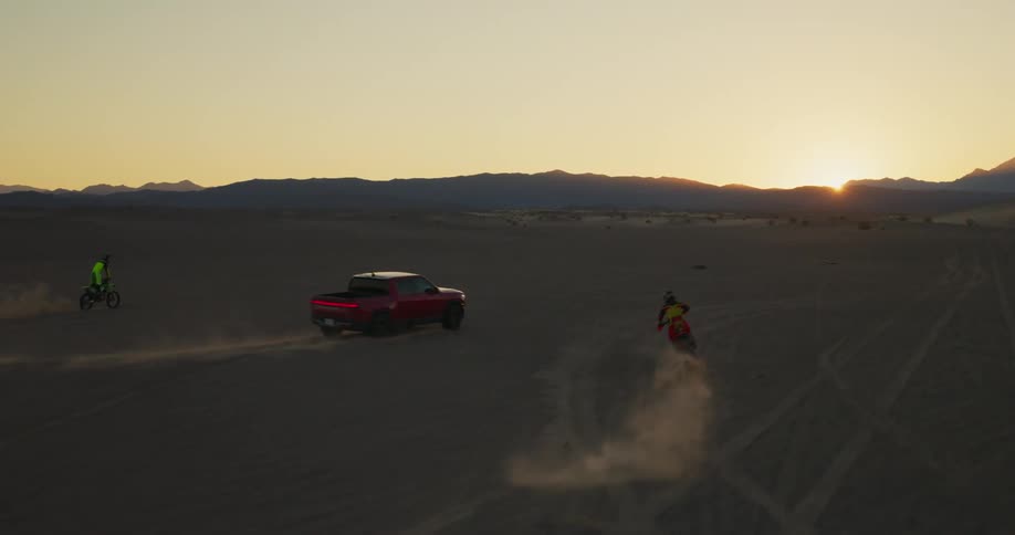 A red pickup truck and two motorcycles doing motocross next to them and everyone is riding in a sandy desert