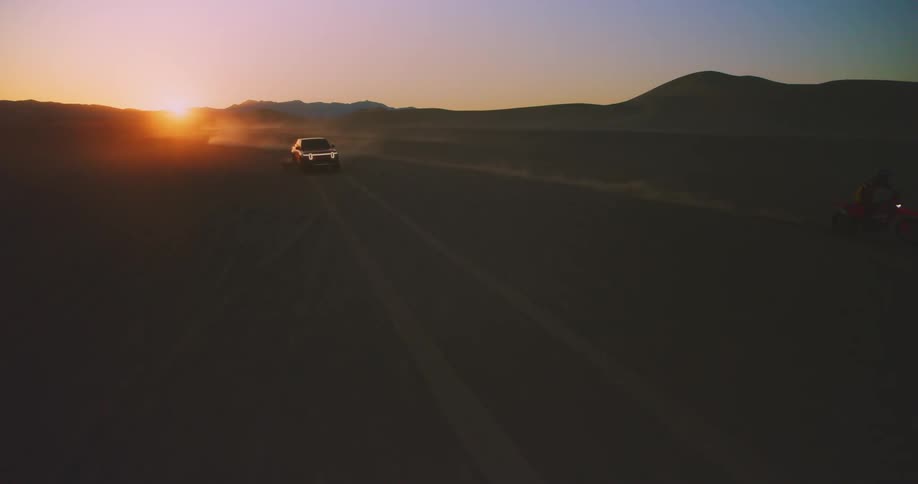 Two motorbikes and a pickup truck driving in a desert with sunset light in the background