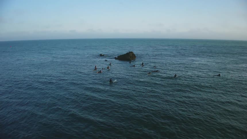 Several surfers next to a rock on the surface of the water waiting for waves