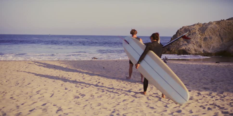 Three surfers walking across the sand and towards the ocean talking and smiling while carrying their surfboards